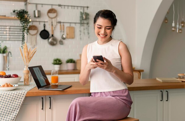 Smiling young asian woman using mobile phone while sitting in kitchen room at home with laptop