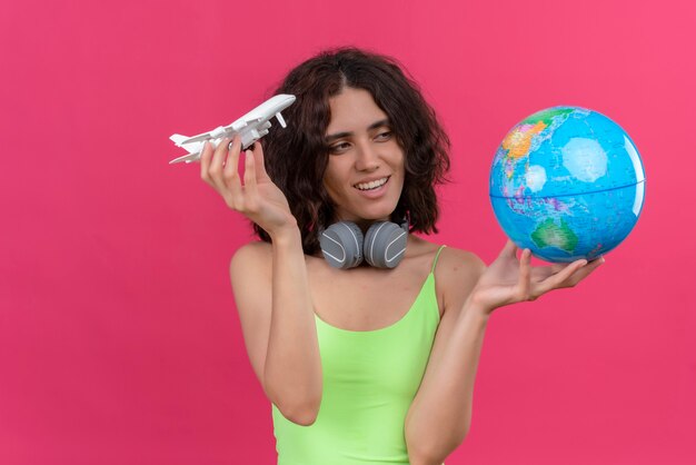 Улыбающаяся молодая привлекательная женщина с короткими волосами в зеленом топе в наушниках держит игрушечный самолет и смотрит на глобус