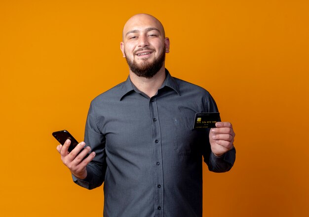 Smiling young bald call center man holding credit card and mobile phone isolated on orange wall