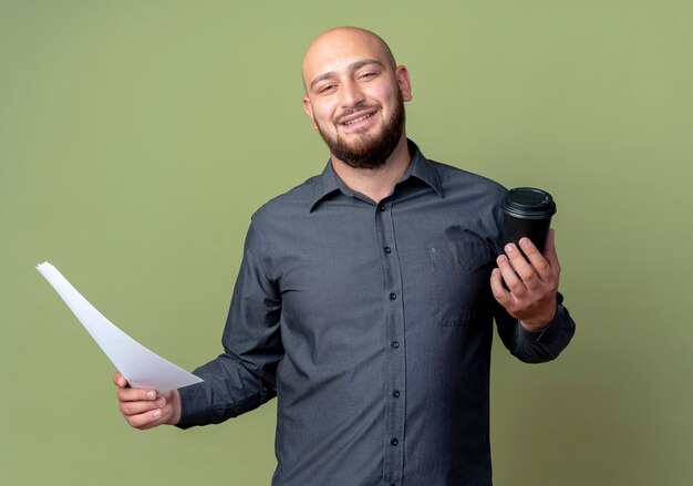 Smiling young bald call center man holding document and plastic coffee cup isolated on olive green wall