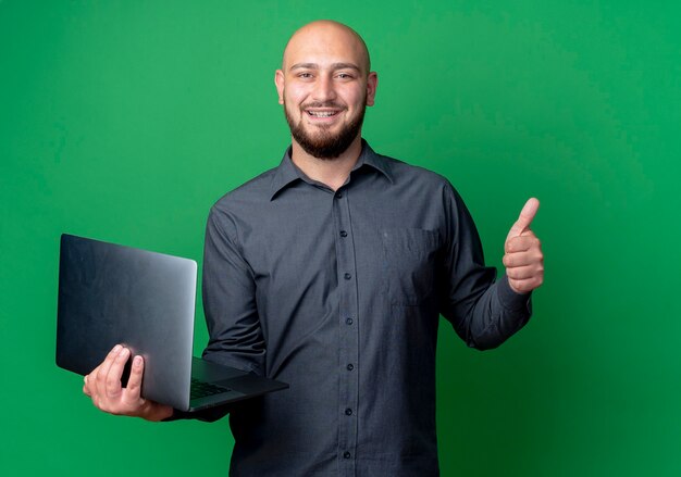 Smiling young bald call center man holding laptop and showing thumb up isolated on green wall