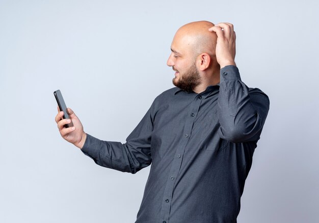Улыбающийся молодой лысый человек из колл-центра, держащий и смотрящий на мобильный телефон с рукой на голове, изолирован на белой стене