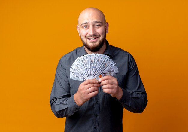 Smiling young bald call center man holding money isolated on orange wall