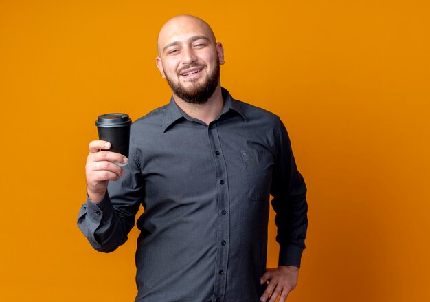 Smiling young bald call center man holding plastic coffee cup and putting hand on waist isolated on orange wall