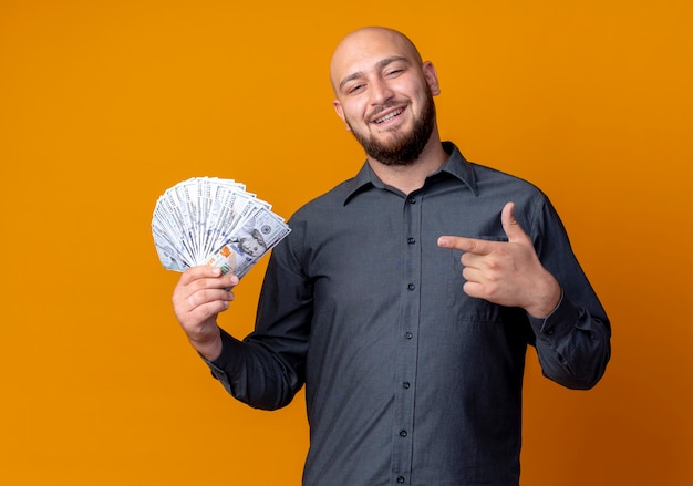 Smiling young bald call center man holding and pointing at money isolated on orange wall