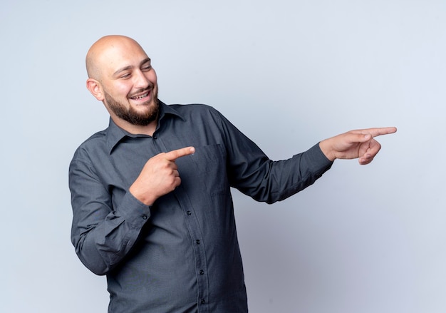 Smiling young bald call center man looking and doing you gesture at side isolated on white wall