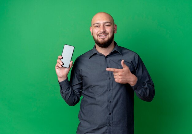 Smiling young bald call center man showing and pointing at mobile phone isolated on green wall