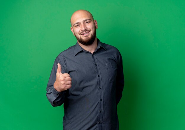 Smiling young bald call center man showing thumb up isolated on green wall