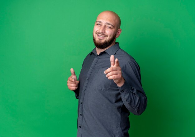 Smiling young bald call center man standing in profile view and doing you gesture at front isolated on green wall