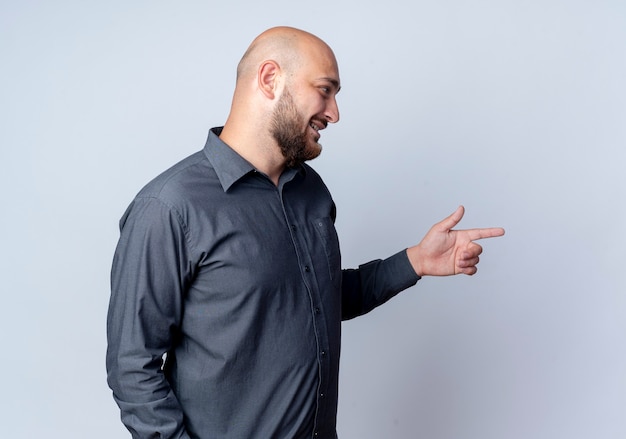 Smiling young bald call center man standing in profile view looking and pointing straight isolated on white wall