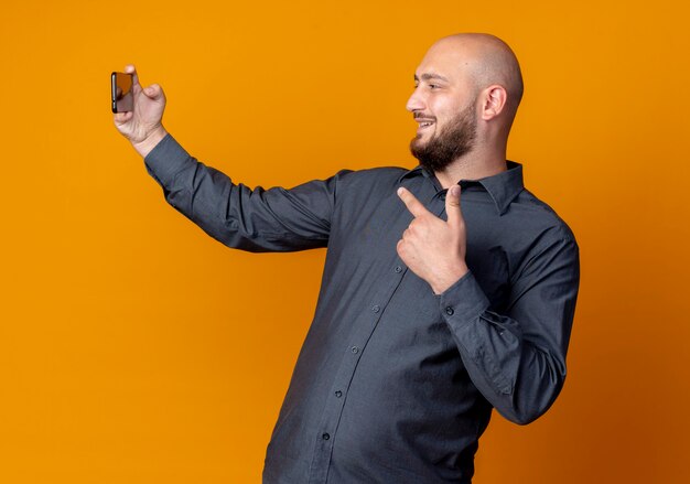 Smiling young bald call center man taking selfie and pointing at mobile phone isolated on orange wall