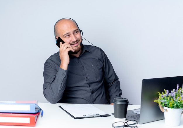 작업 도구가 측면을보고 흰 벽에 고립 된 전화 통화와 책상에 앉아 헤드셋을 착용하는 젊은 대머리 콜 센터 남자 미소