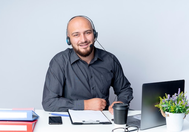 Sorridente giovane calvo call center uomo che indossa la cuffia avricolare seduto alla scrivania con strumenti di lavoro mettendo le mani sulla scrivania isolato sul muro bianco