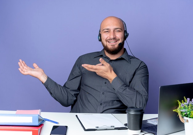 Smiling young bald call center man wearing headset sitting at desk with work tools showing empty hand and pointing at it isolated on purple wall