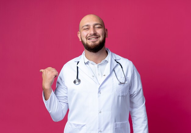 Smiling young bald male doctor wearing medical robe and stethoscope points at side isolated on pink with copy space