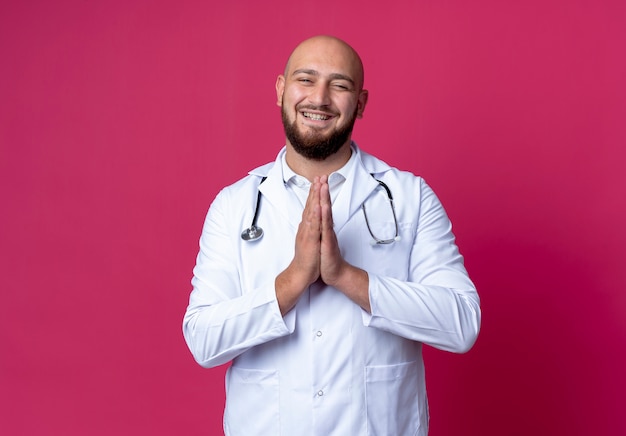 Sorridente giovane medico maschio calvo che indossa abito medico e stetoscopio che mostra pregare gesto isolato sul rosa