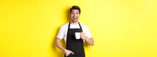 노란색 배경 위에 서 있는 커피 컵을 들고 검은 앞치마를 입은 웃고 있는 젊은 바리스타