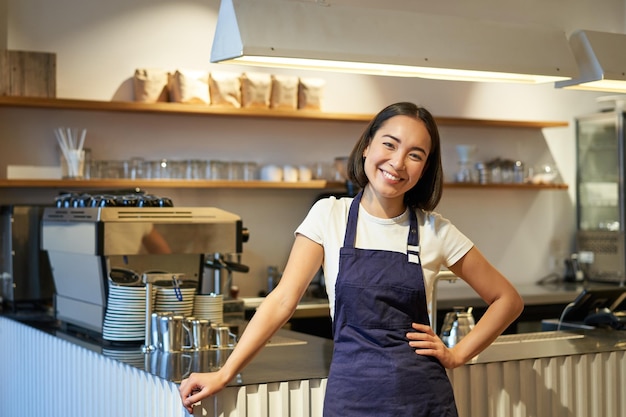 Smiling young barista wearing apron working in cafe standing near counter with coffee machine lookin