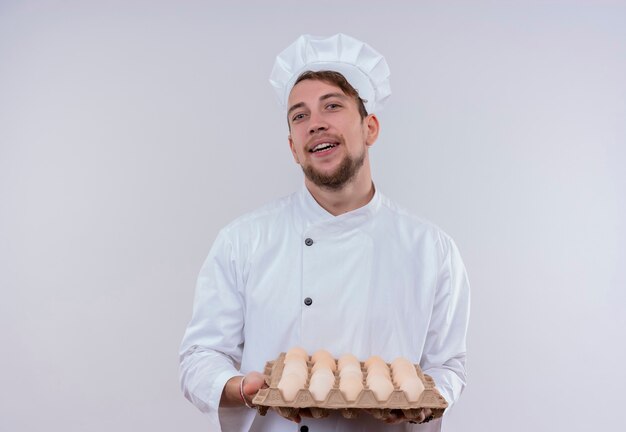 A smiling young bearded chef man wearing white cooker uniform and hat holding a carton of organic eggs while looking on a white wall