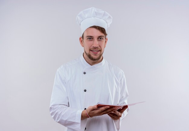 A smiling young bearded chef man wearing white cooker uniform and hat holding notebook while looking on a white wall