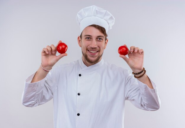 Улыбающийся молодой бородатый повар в белой униформе и шляпе держит красные помидоры, глядя на белую стену