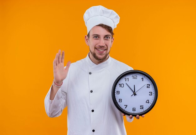 A smiling young bearded chef man in white uniform holding wall clock while looking on an orange wall