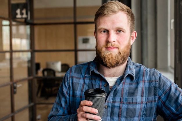 Smiling young bearded man holding disposable coffee cup looking at camera