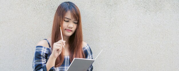 Smiling young beautiful asian woman thinking with writing book on concrete wall background. Vintage effect style pictures.