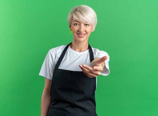 Smiling young beautiful female barber in uniform holding out hand at camera isolated on green background