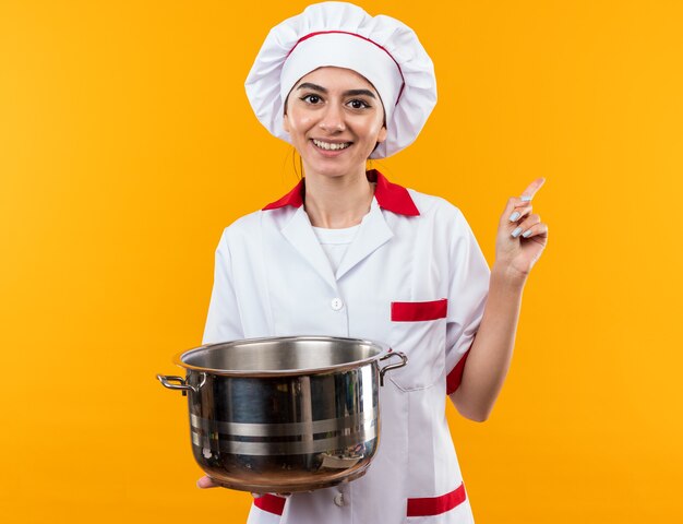 Smiling young beautiful girl in chef uniform holding and looking at saucepan points at side 