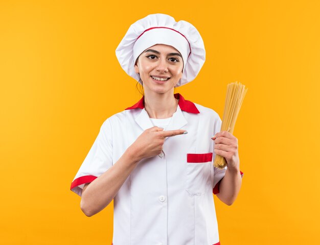 Улыбающаяся молодая красивая девушка в униформе шеф-повара держит и указывает на спагетти