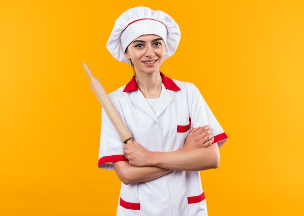 Sorridente giovane bella ragazza in uniforme da chef tenendo il perno del rullo attraversando le mani isolate sulla parete arancione