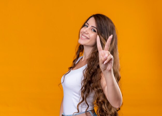 Smiling young beautiful girl doing peace sign standing in profile view on isolated orange space with copy space