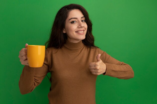 차 한잔 들고 엄지 손가락을 보여주는 젊은 아름 다운 소녀 미소는 녹색 벽에 고립