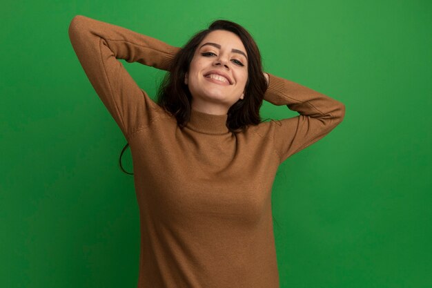 Smiling  young beautiful girl holding hands behind head isolated on green wall