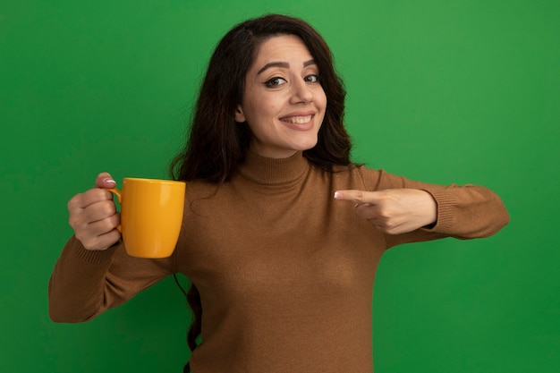 Улыбающаяся молодая красивая девушка держит и указывает на чашку чая, изолированную на зеленой стене