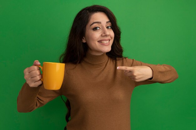 Smiling young beautiful girl holding and points at cup of tea isolated on green wall