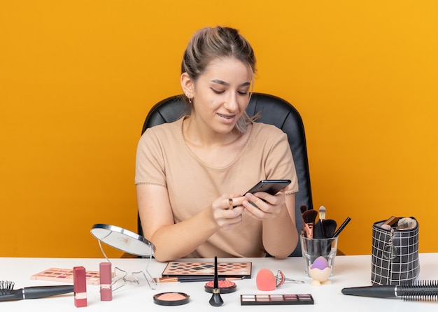 Free photo smiling young beautiful girl sits at table with makeup tools holding makeup brush and looking at phone in her hand isolated on orange background