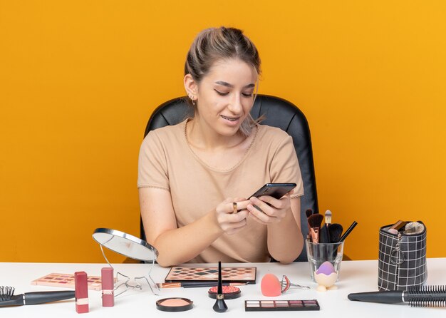 Smiling young beautiful girl sits at table with makeup tools holding makeup brush and looking at phone in her hand isolated on orange background