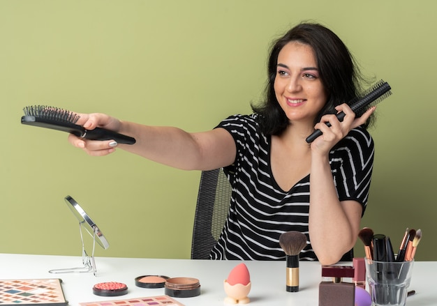 Smiling young beautiful girl sits at table with makeup tools holding out combs at side isolated on olive green background