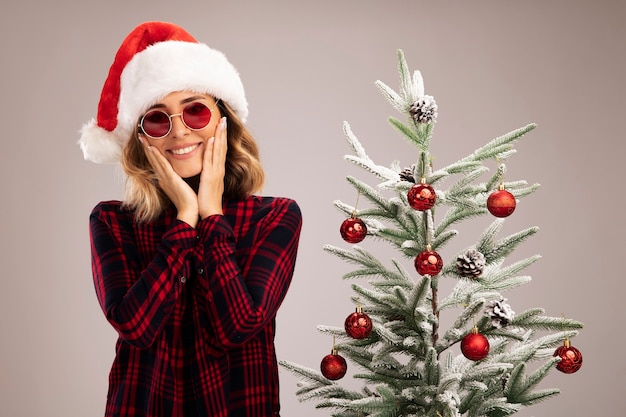 Free photo smiling young beautiful girl standing nearby christmas tree wearing christmas hat with glasses covered cheeks with hands isolated on white background