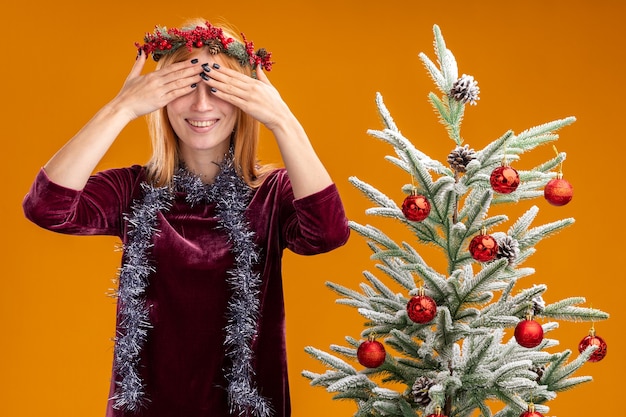 Foto gratuita sorridente giovane bella ragazza in piedi vicino albero di natale che indossa abito rosso e corona con ghirlanda sul collo coperto gli occhi con le mani isolate sulla parete arancione