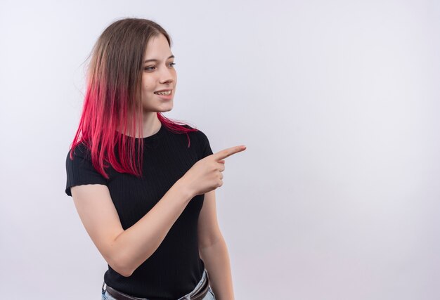 Smiling young beautiful girl wearing black t-shirt points finger to side on isolated white background with copy space