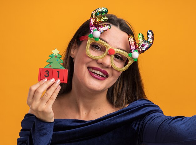 Улыбающаяся молодая красивая девушка в синем платье и рождественских очках держит игрушку и камеру на оранжевом фоне