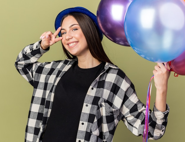 Foto gratuita sorridente giovane bella ragazza che indossa un cappello blu con palloncini che mostrano gesto di pace