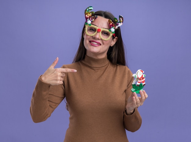 smiling young beautiful girl wearing brown sweater with christmas glasses holding and points at toy isolated on blue wall