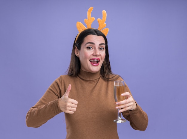 青い背景で隔離の親指を示すシャンパンのガラスを保持しているクリスマスの髪のフープと茶色のセーターを着て笑顔の若い美しい少女