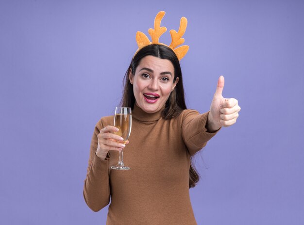 Улыбающаяся молодая красивая девушка в коричневом свитере с рождественским обручем для волос, держащая бокал шампанского, показывает палец вверх, изолированные на синем фоне