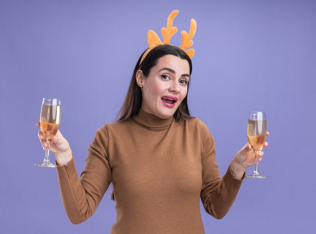 smiling young beautiful girl wearing brown sweater with christmas hair hoop holding two glass of champagne isolated on blue wall