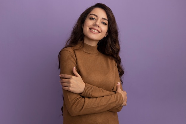 Smiling young beautiful girl wearing brown turtleneck sweater crossing hands isolated on purple wall with copy space